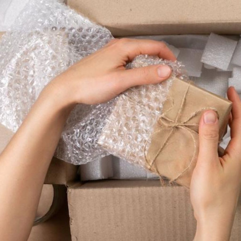 Hands wrapping a small, brown paper-wrapped gift with bubble wrap. The package is tied with a twine bow. The gift is being placed into a cardboard box filled with packing materials.