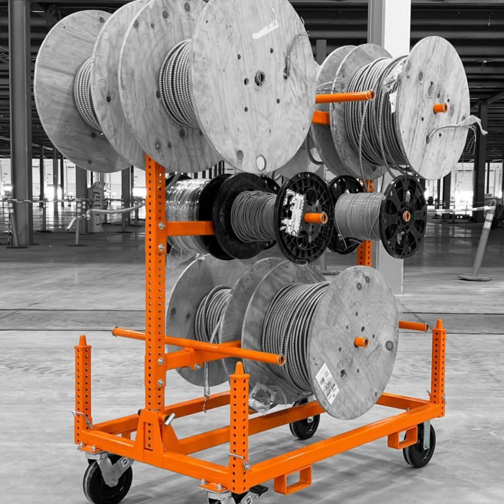 An industrial orange cart holds multiple large spools of cable in a warehouse setting. The spools are both wooden and plastic, with cables coiled and neatly wrapped around them. The cart has wheels for mobility.