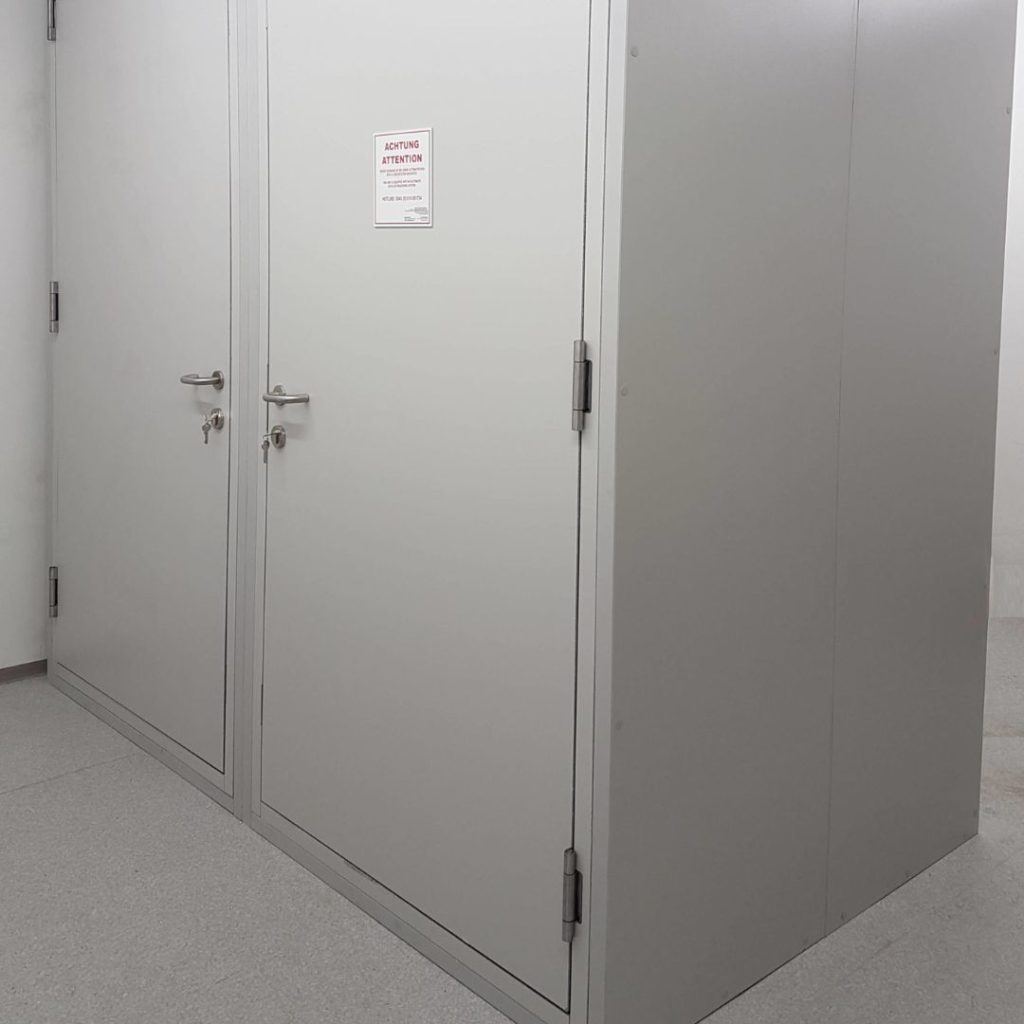 A white office storage room with two closed metal doors, each with a handle. Above the handles, a small notice is posted. The floor is light gray, and the walls are plain white. The room appears empty and utilitarian.