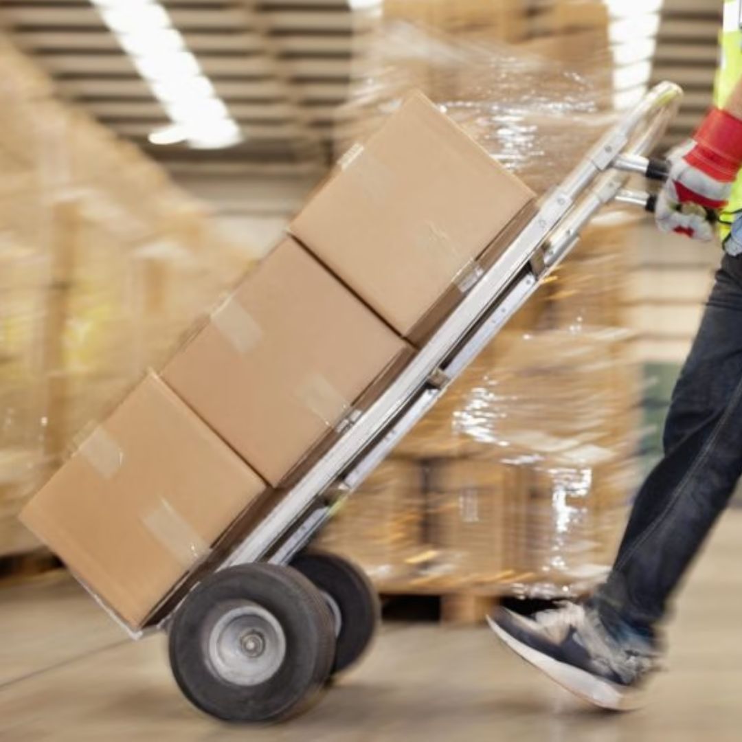 Worker moving stacked cartons on a hand truck through a warehouse aisle