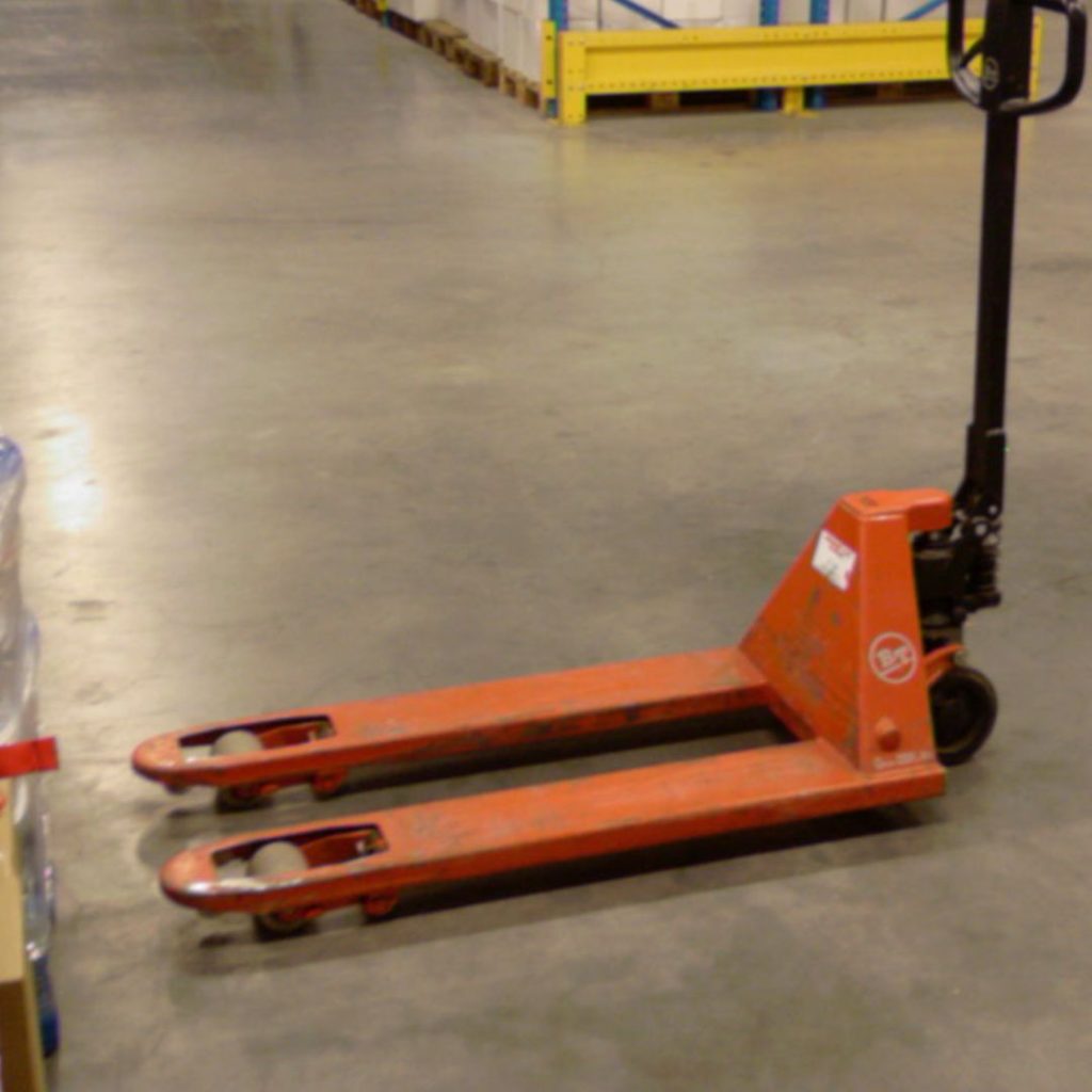 A red pallet jack is positioned on a warehouse floor, surrounded by stacks of boxes and shelves. The jack has a black handle and is ready for moving pallets.