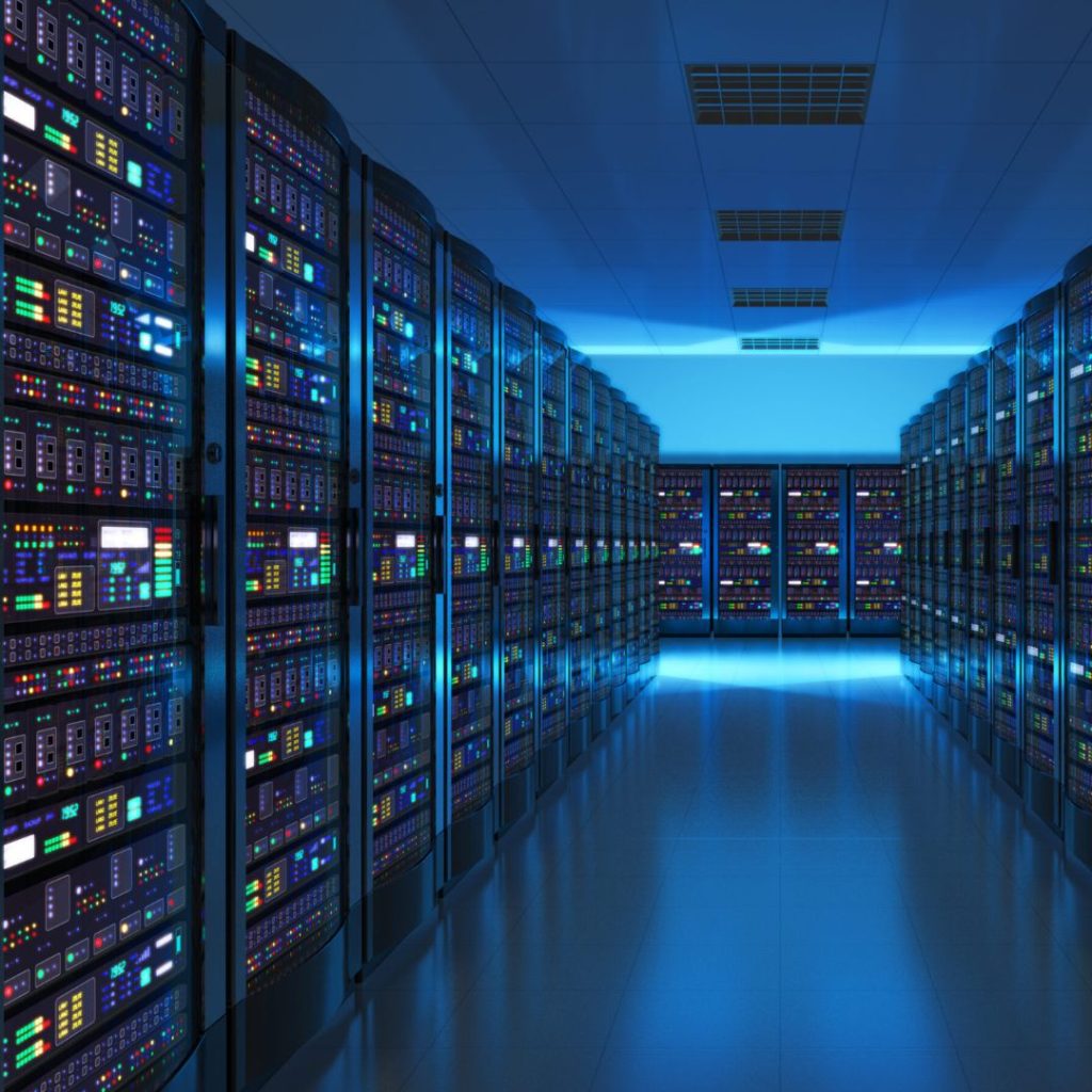 A brightly lit server room with two rows of server racks filled with colorful lights and switches. The room has a reflective floor and a ceiling with panels, giving a high-tech, futuristic appearance.