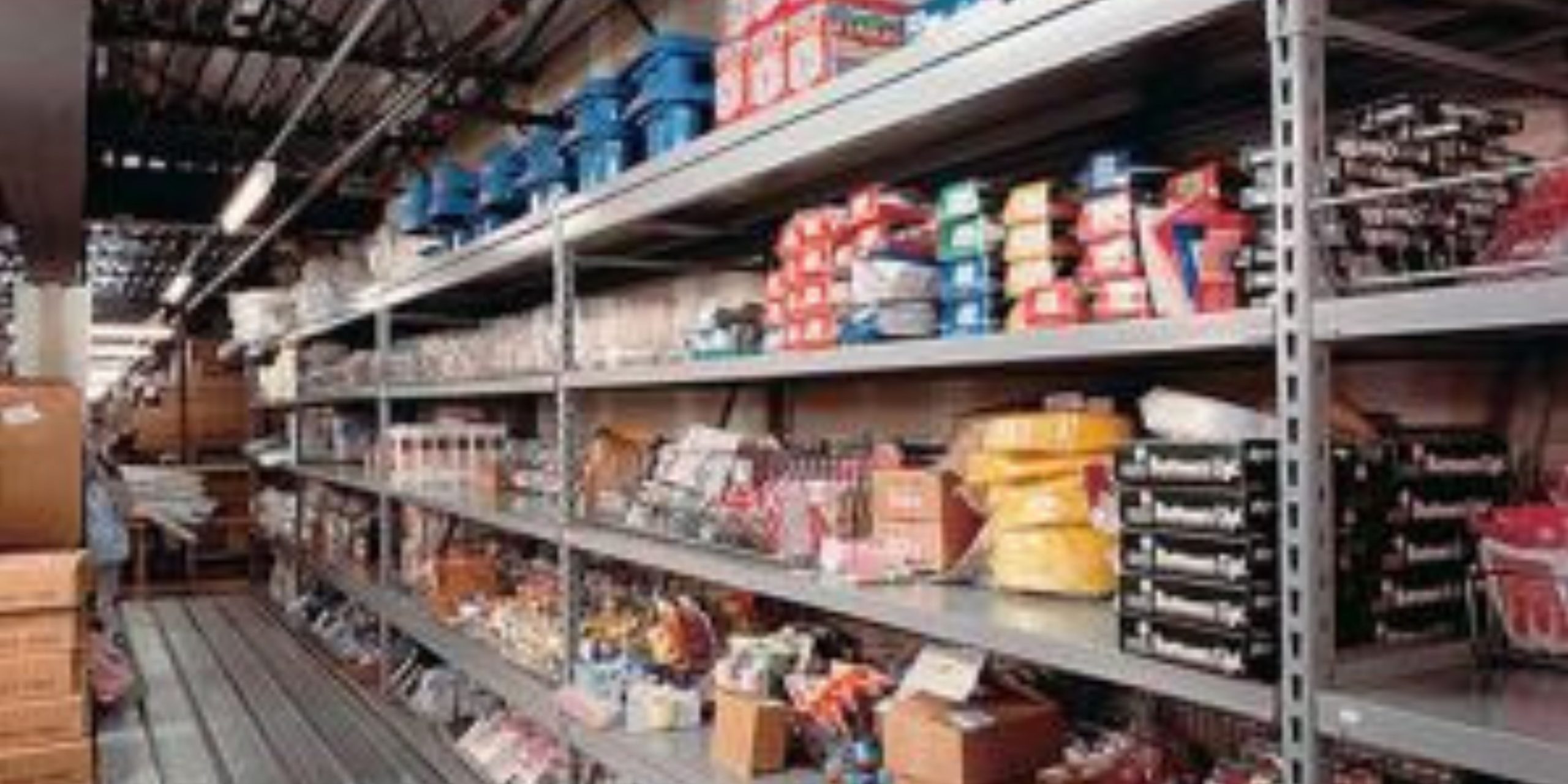 Metal shelves filled with various supplies—including boxes, plastic containers, and packaged goods—are neatly organized in a storeroom or warehouse with tailored shelving solutions, featuring a visible walkway and overhead pipes.