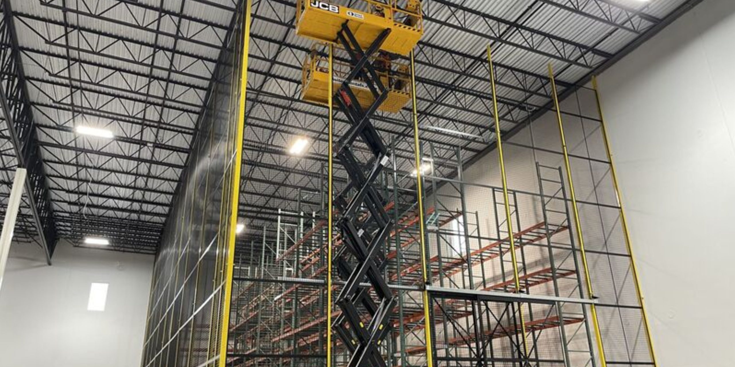 A large indoor warehouse with tall metal racks and a yellow scissor lift raised high, with two workers standing on the lift platform, working near the ceiling.