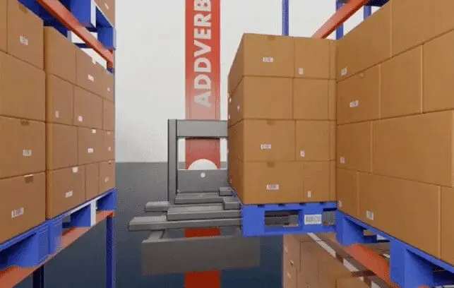 Crane-based ASRS lifting a pallet of cardboard boxes between tall racking aisles in a warehouse.