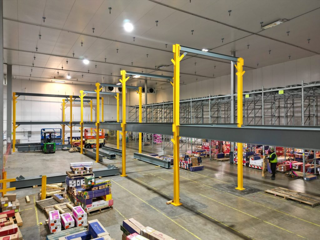 A large warehouse with tall yellow and gray metal shelving under construction, boxes and pallets organized on the floor, and workers in safety vests handling materials and equipment.