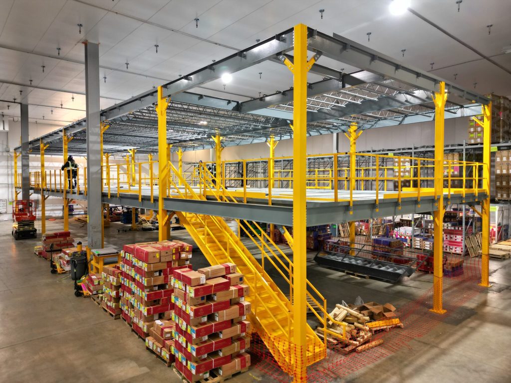 A warehouse interior shows a large yellow metal mezzanine with stairs, surrounded by stacked boxes and pallets. Bright lighting highlights workers and construction materials scattered throughout the space.