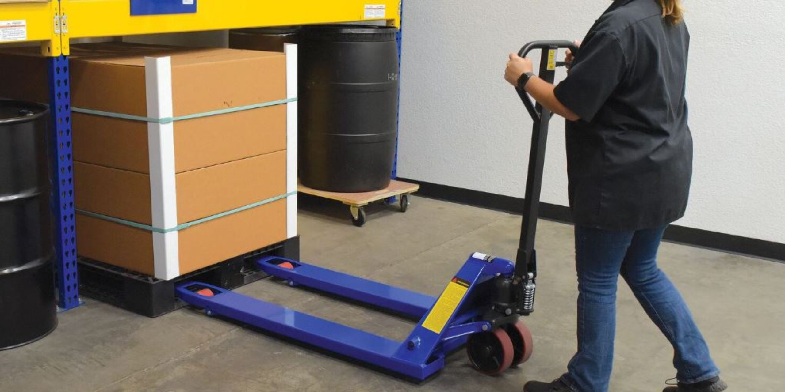 Worker operating a manual pallet truck beside pallet rack in a warehouse