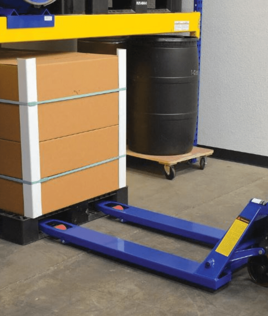 A blue pallet jack positioned to lift a stacked pallet of brown cardboard boxes secured with white straps in an industrial warehouse setting. A black barrel is visible in the background.