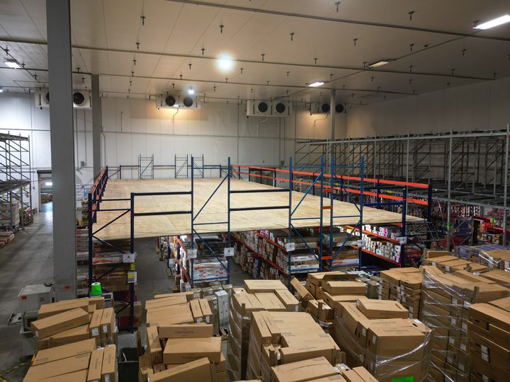 Warehouse interior with stacked cardboard boxes in the foreground, tall metal racks with goods, and an elevated empty platform structure in the center. The space is brightly lit, with a high ceiling and visible ventilation units.