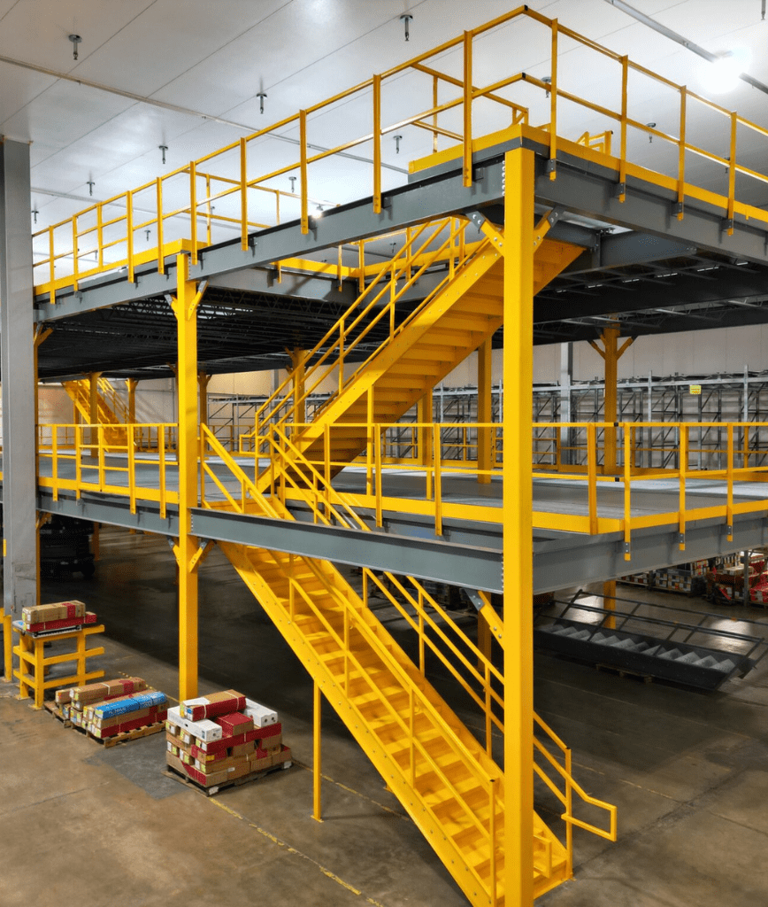 A large industrial warehouse features a bright yellow metal mezzanine structure with stairs and railings, providing an elevated walkway and storage area above the main floor, where pallets of goods are stacked.