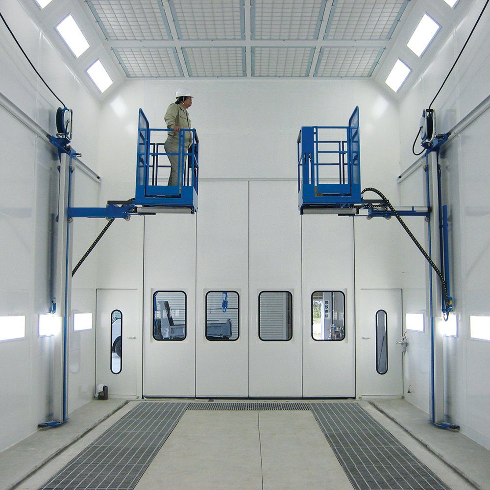 A person in safety gear stands on a blue hydraulic lift inside a large, brightly-lit industrial room with white walls, high ceilings, and large doors. Another identical lift is positioned on the opposite wall.