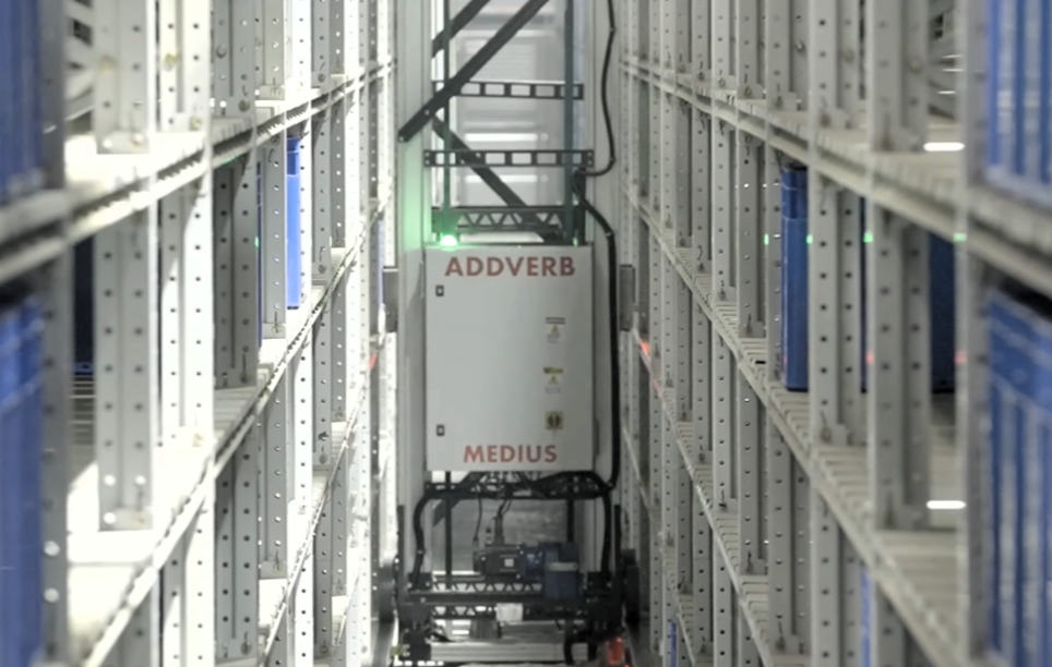 A robotic warehouse system labeled ADDVERB MEDIUS moves between tall shelves filled with blue bins, navigating a narrow aisle in an automated storage facility.