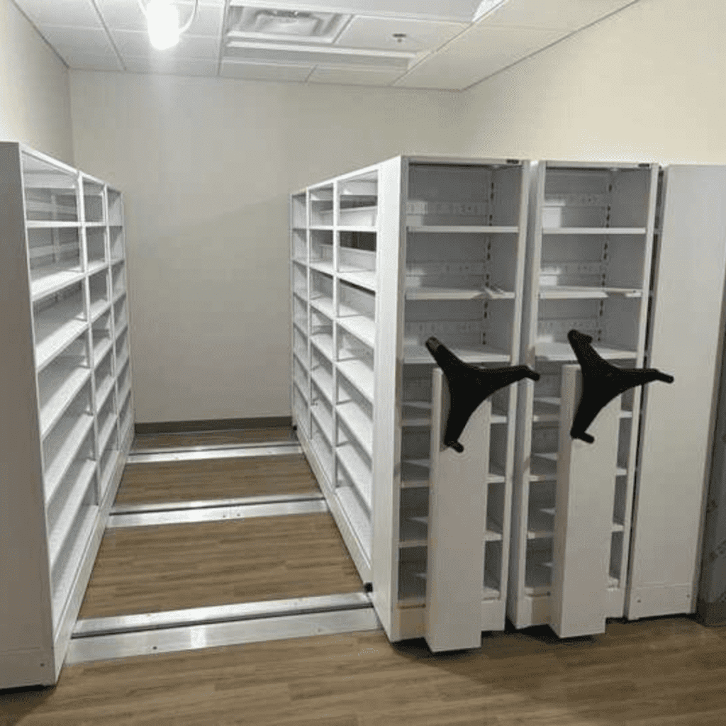 White sliding mobile shelving units with empty shelves are positioned on tracks over a wooden floor in a room with a white ceiling and fluorescent lighting. Two shelves have large black handles for moving the units.