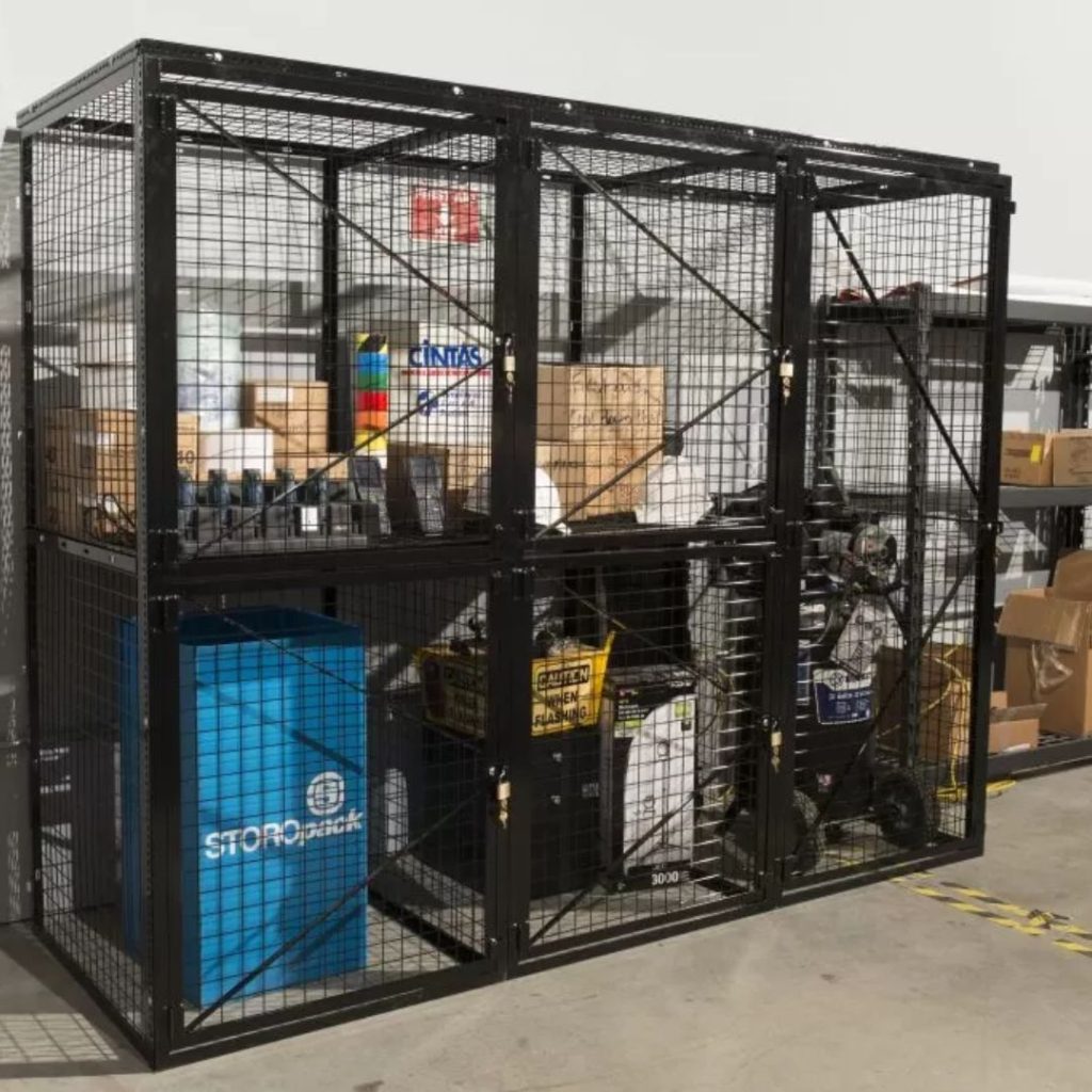 A large black metal storage cage filled with boxes, equipment, and supplies sits in a warehouse. The cage is locked and placed against a white wall with shelves and more boxes surrounding it.