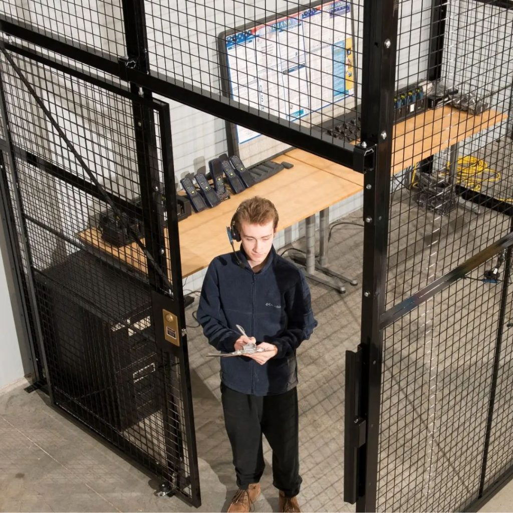 A person stands in a secure, caged area with a clipboard, wearing a headset. Behind them is a workbench with tools and equipment, and a whiteboard attached to the wall. The area appears industrial and organized.