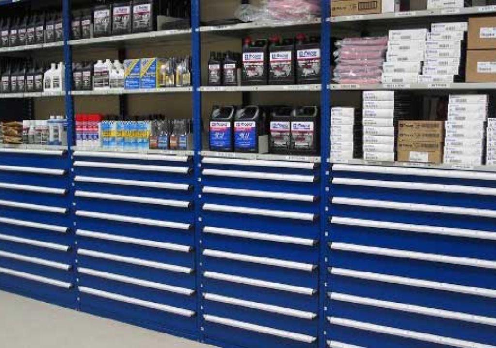Shelves stocked with automotive supplies, including oil containers, small boxes, and bottles, sit above rows of closed blue drawers with white handles, featuring Equipto and Borroughs Shelving ideal for part shelving in a workshop or garage setting.