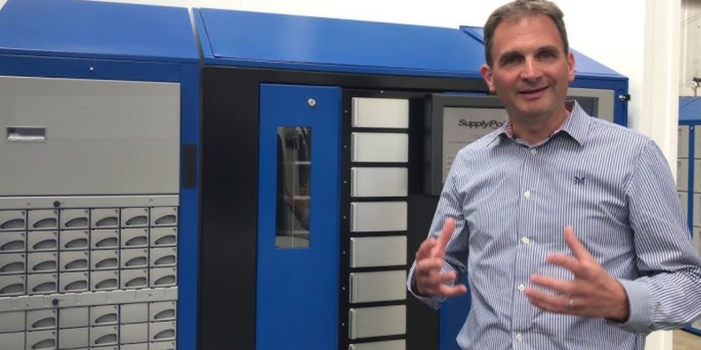 A man in a striped shirt stands in front of a large blue and gray industrial machine with multiple compartments, gesturing with his hands while speaking.