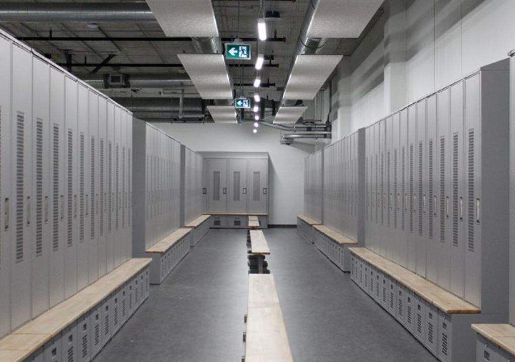 A modern locker room with rows of gray metal lockers on both sides, wooden benches in the center, and ceiling lights and ventilation ducts overhead. The room is empty and clean.