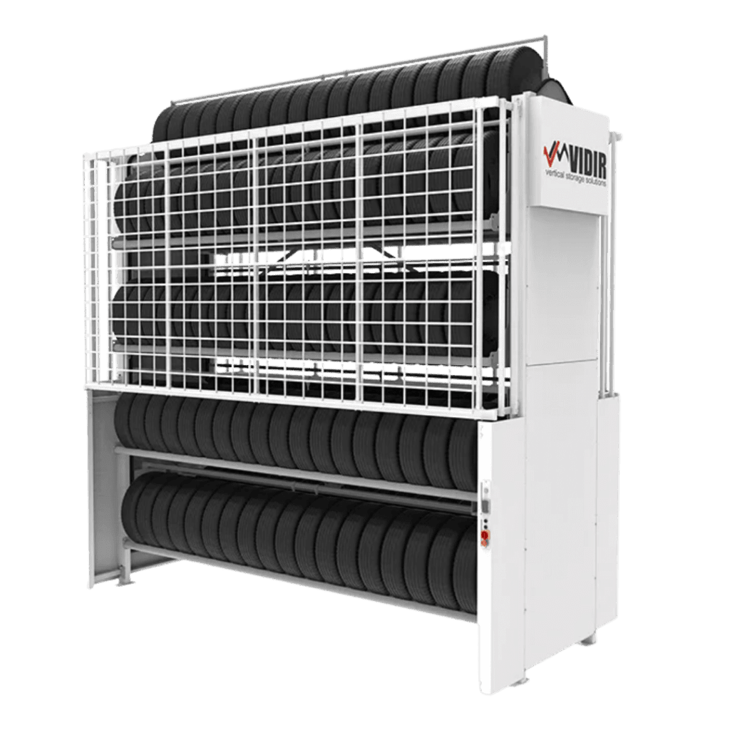 A vertical tire carousel storage system by Vidir Vertical Storage Solutions holds multiple rows of black tires, enclosed in a white metal frame with a mesh safety guard and labeled VIDIR on the side.