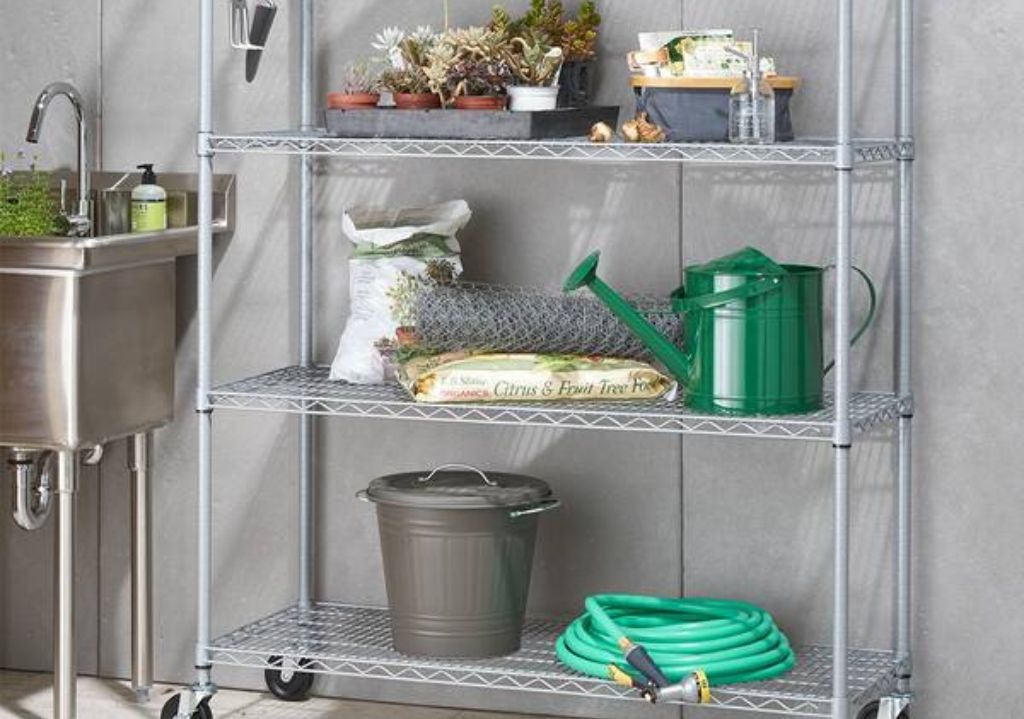 A Heavy Duty Parts Shelving unit on wheels holds potted plants, gardening supplies, a green watering can, a gray bucket, a garden hose, wire fencing, and bags of soil. A sink is beside the shelves.