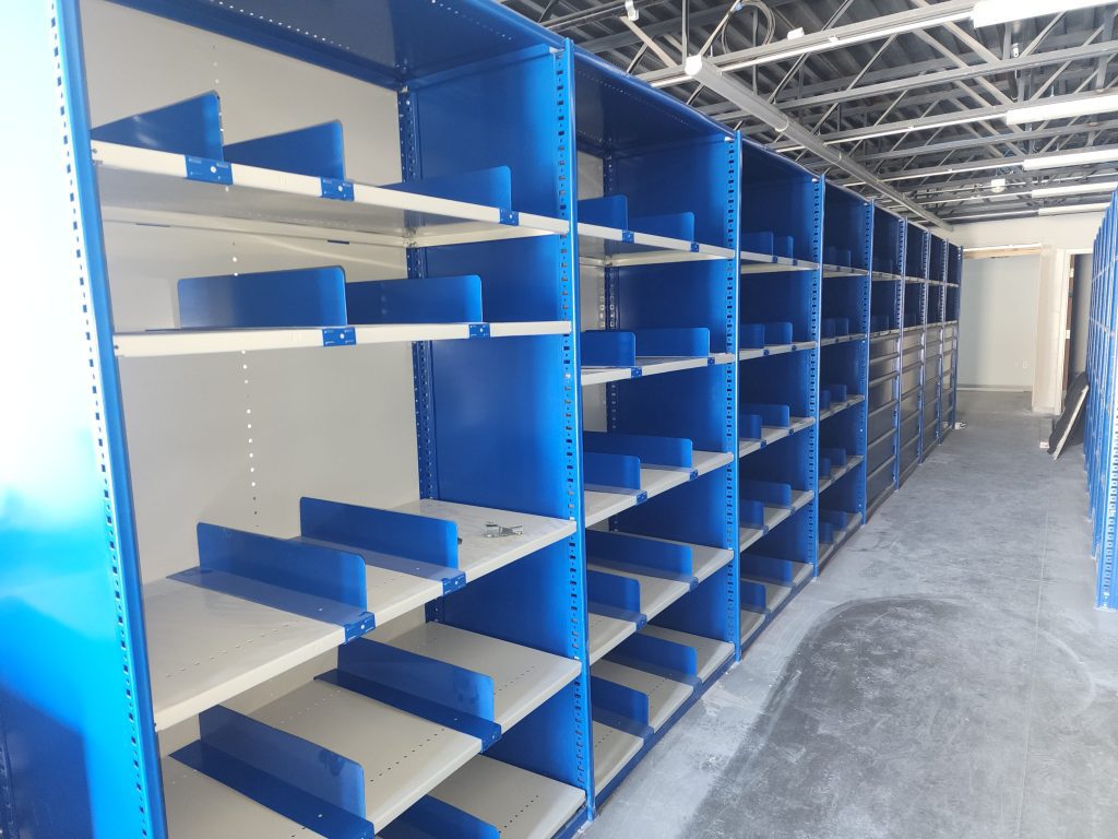 Rows of empty blue metal shelving units inside a brightly lit, unfinished room with a visible ceiling structure and bare concrete floor.