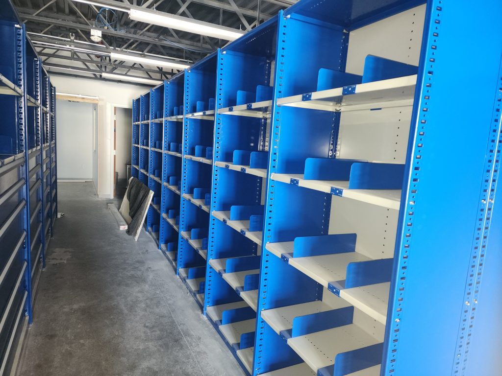 Rows of empty blue metal shelves line both sides of a hallway in a brightly lit room with unfinished ceiling and exposed pipes. Some shelf panels and materials rest on the concrete floor.