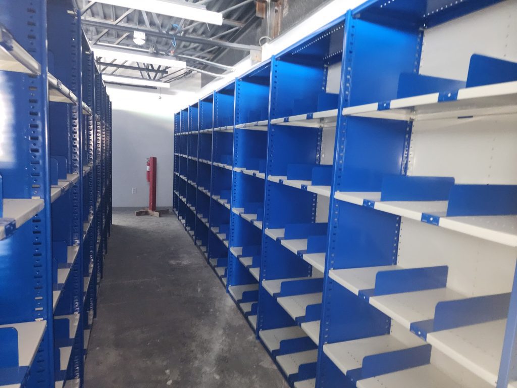 Rows of empty blue metal shelves line both sides of a concrete floor aisle in a well-lit industrial or storage room with exposed ceiling beams.