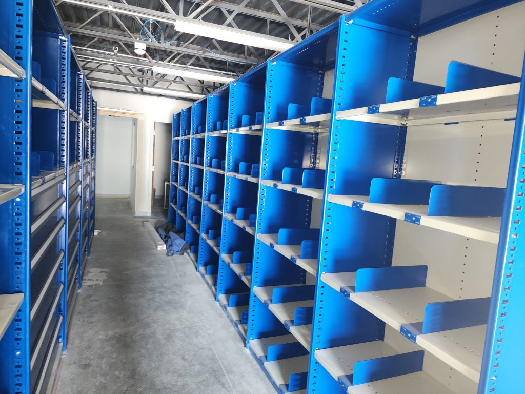 Rows of empty blue metal shelves line both sides of a narrow aisle in a well-lit storage or warehouse area, with a concrete floor and exposed ceiling beams visible above.