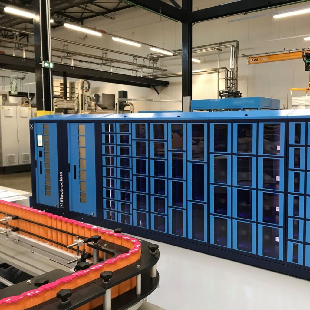 A large, blue automated storage and retrieval system with multiple compartments stands in an industrial facility, surrounded by machinery and exposed pipes. In the foreground, a motorized conveyor with orange and pink segments is visible.
