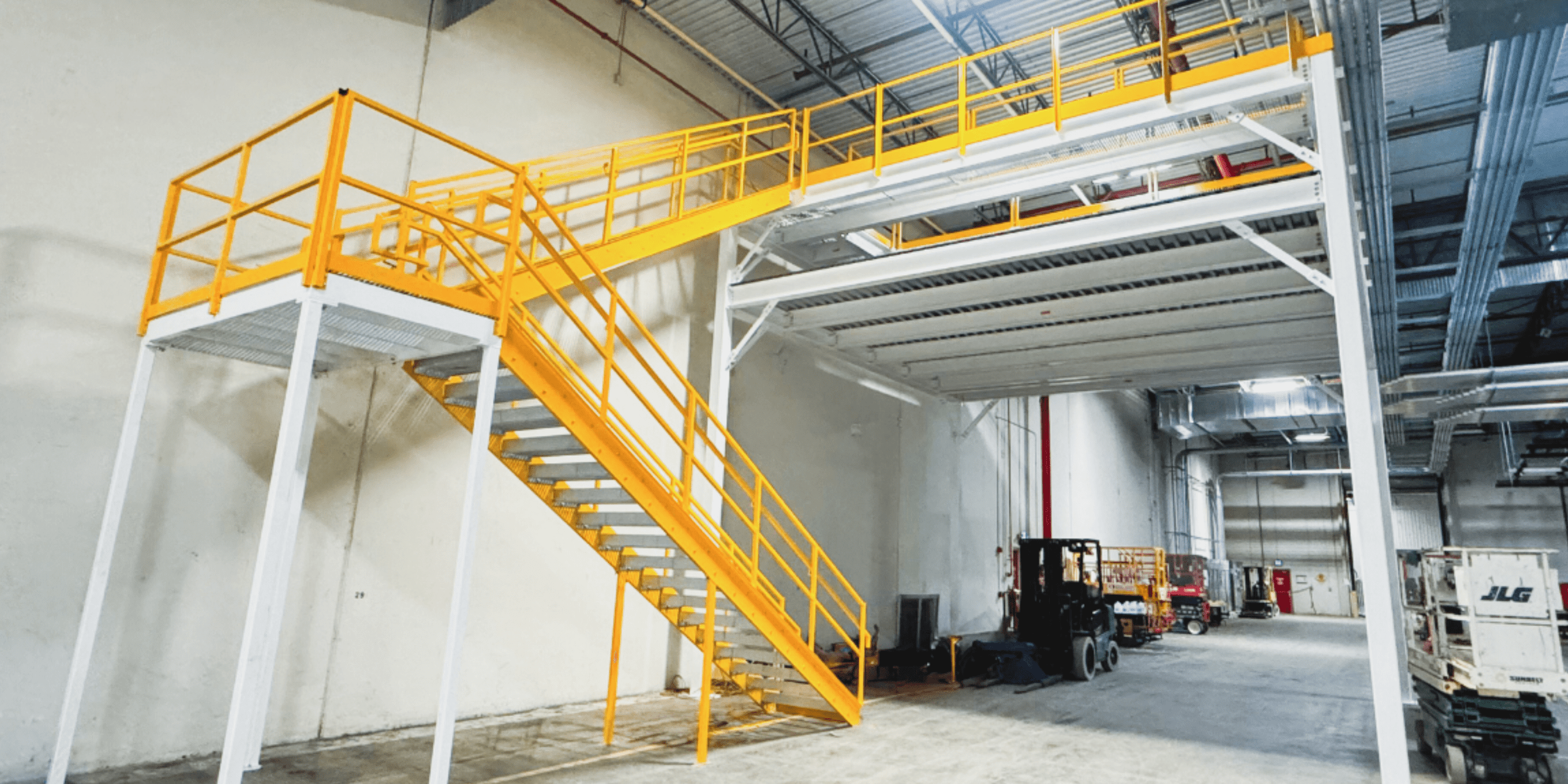 A large industrial warehouse with a yellow metal staircase and railing leading to an elevated white mezzanine platform. Equipment and storage items are visible in the background.