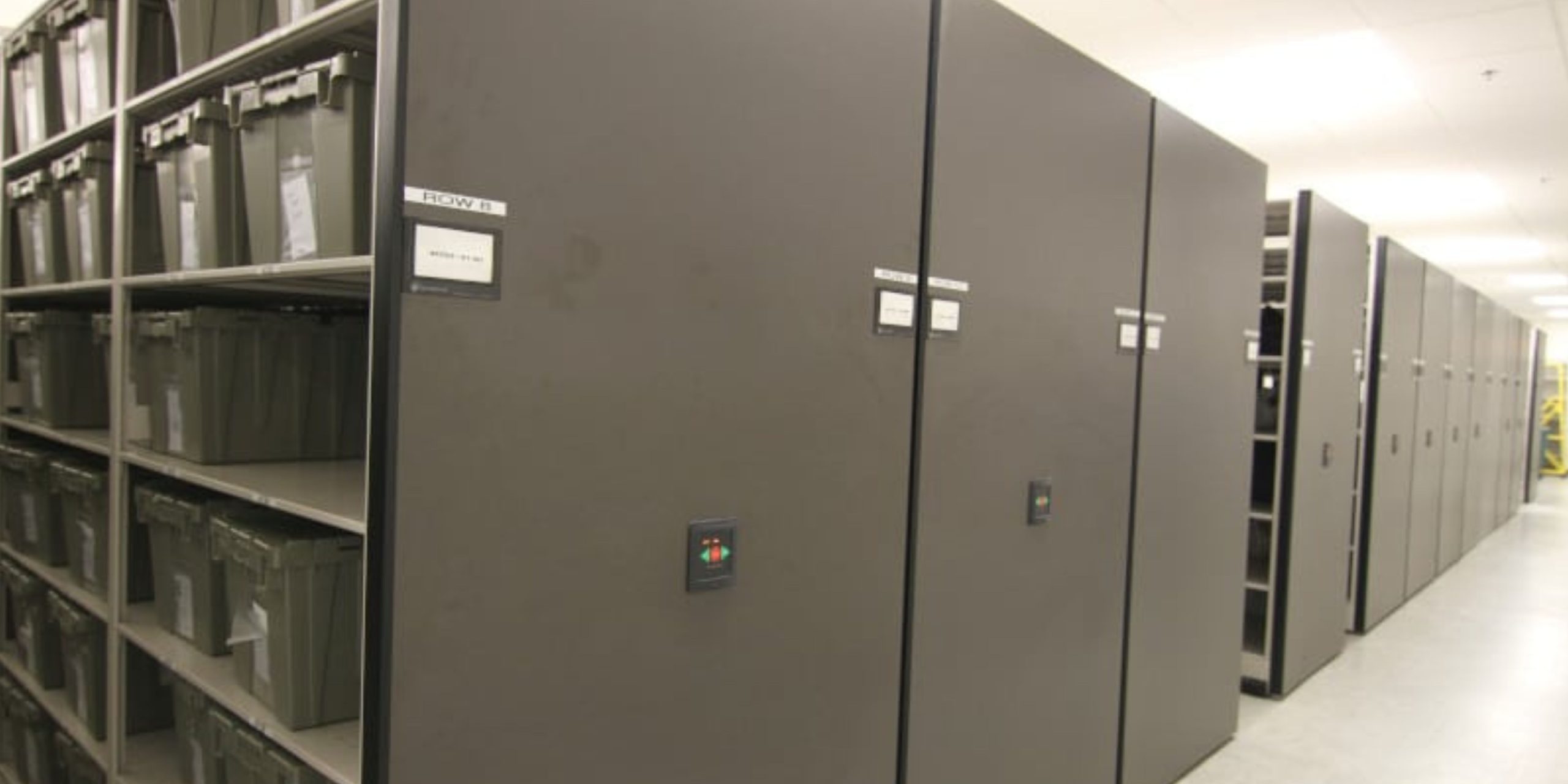 High density shelving ranges with end-panel controls storing labelled cases and cartons in a government storage room