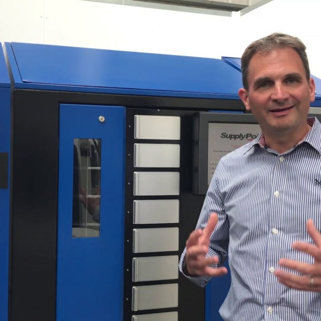 A man in a striped shirt gestures with his hands while standing next to a large blue industrial machine with multiple compartments and a SupplyPoint label.