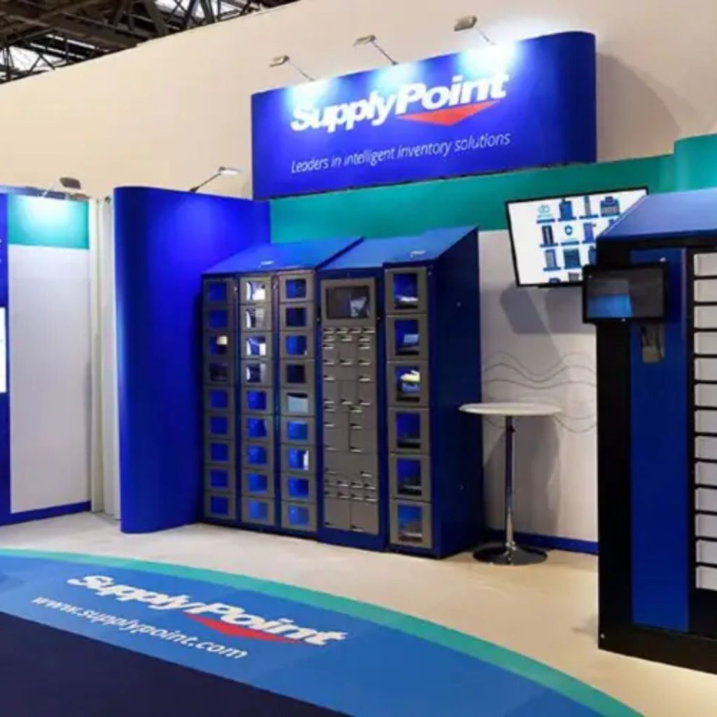 A trade show booth for SupplyPoint displays blue and gray automated inventory storage lockers, a digital screen showing interface graphics, and a branded floor mat with the logo and slogan Leaders in intelligent inventory solutions.