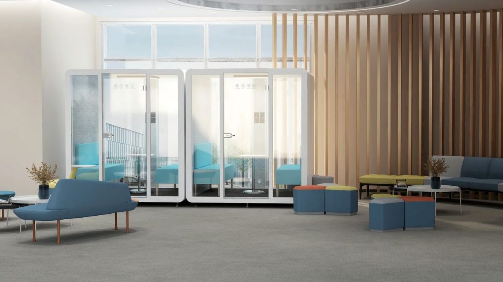 A modern office lounge with blue seating, colorful ottomans, two glass phone booths, small tables with plants, and a wood slat accent wall, all in a bright space with large windows.