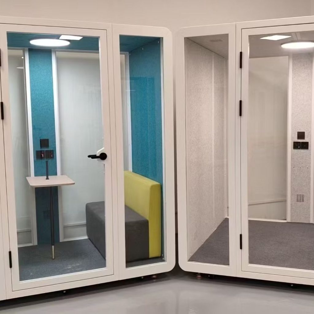 Two modern, glass-walled office pods are shown side by side. One pod has a small table and a yellow cushioned bench, while the other pod is empty, both designed for private work or phone calls.