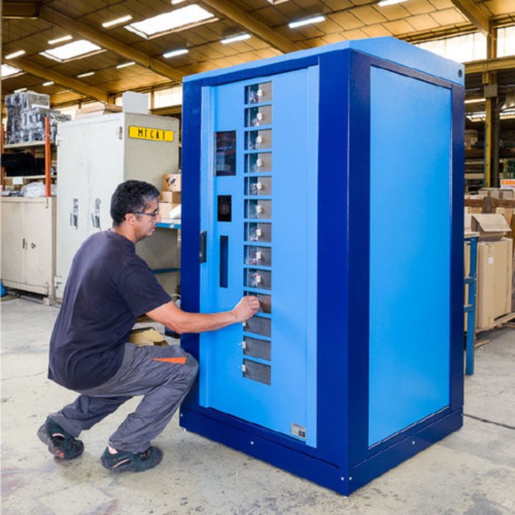A man kneels and operates a large blue industrial vending machine in a warehouse, using buttons on the front panel. Shelves with various items are visible through the machine’s transparent sections.