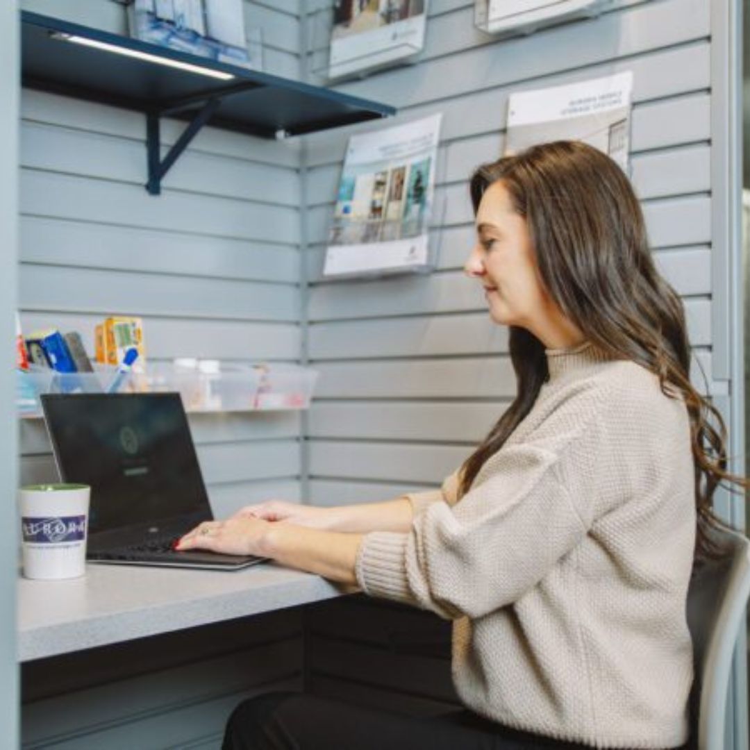 A woman with long brown hair, wearing a beige sweater, sits at a desk typing on a laptop. A coffee mug is nearby, and shelves with brochures and office supplies are visible in the background.