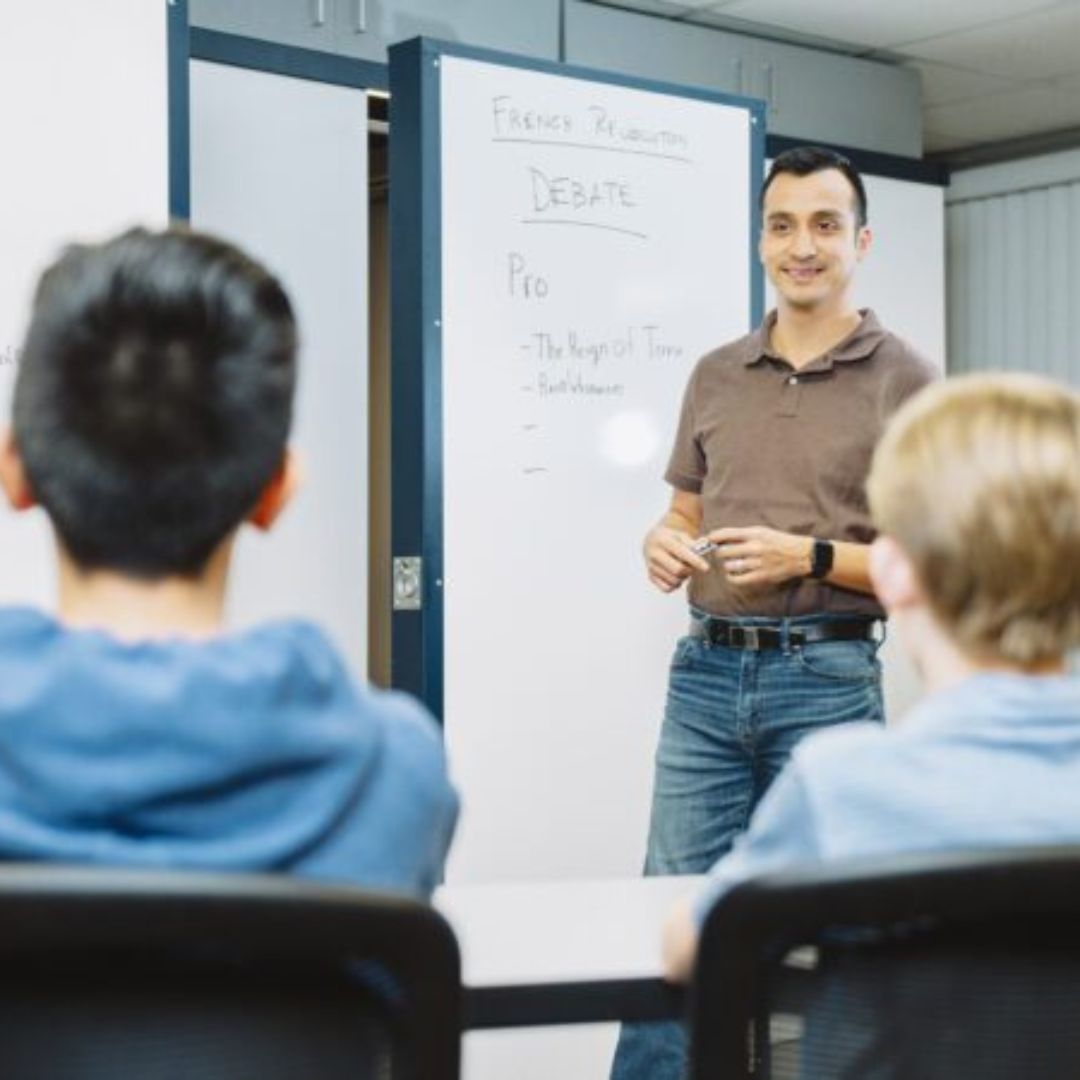 A teacher stands in front of a whiteboard labeled French Revolution Debate and faces two seated students, explaining points listed under Pro while holding a marker and smiling.