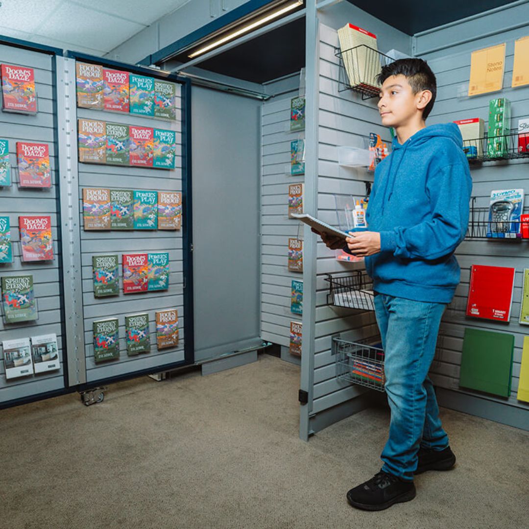 A boy in a blue hoodie holding a tablet stands in a library or bookstore aisle, surrounded by shelves filled with LEGO magazines, books, and stationery items.