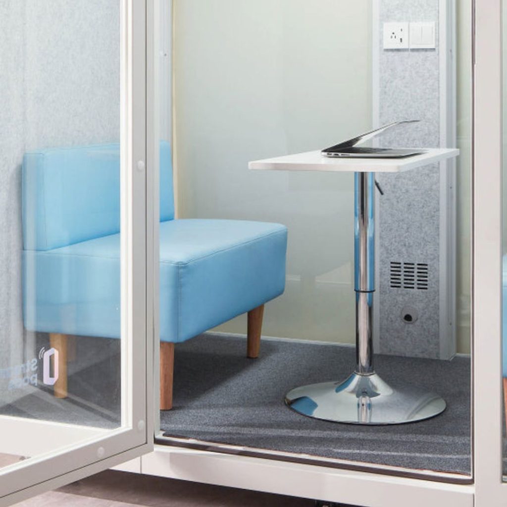 A small, glass-walled booth with a light blue cushioned chair, a white table with a chrome base, and a closed laptop on the table. The space has grey carpeting and power outlets on the wall.