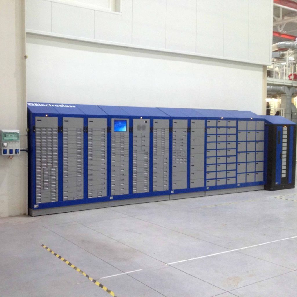 A large blue and gray industrial vending machine with multiple small lockers stands against a wall in a spacious, clean factory or warehouse environment.
