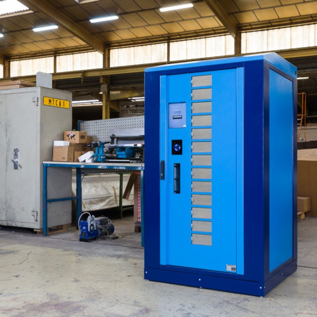 A large blue industrial vending machine stands on a factory floor near a workbench with tools, boxes, and machinery. The background includes shelves, equipment, and a cabinet labeled MICIM.