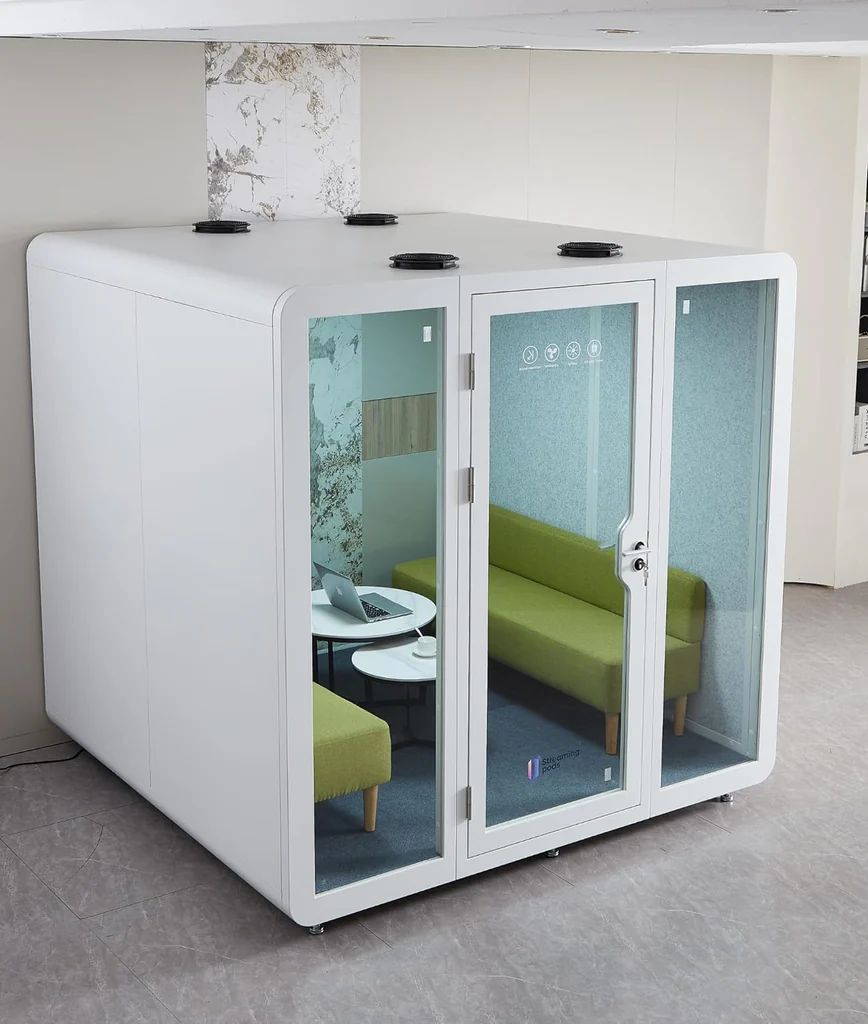 A modern office meeting pod with glass doors and windows, containing two green sofas, a small round table, and power outlets, situated in a bright indoor space with tiled flooring.
