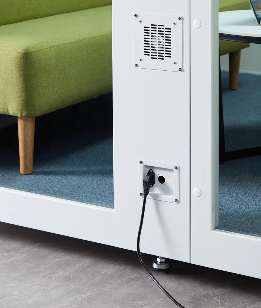 A black electrical plug is connected to an outlet on a white partition wall with vents, next to a green sofa with wooden legs and a blue carpeted floor.