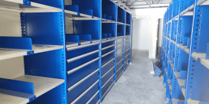 A room with rows of empty blue metal shelving units and drawers on a gray concrete floor, with a partially constructed doorway visible in the background.