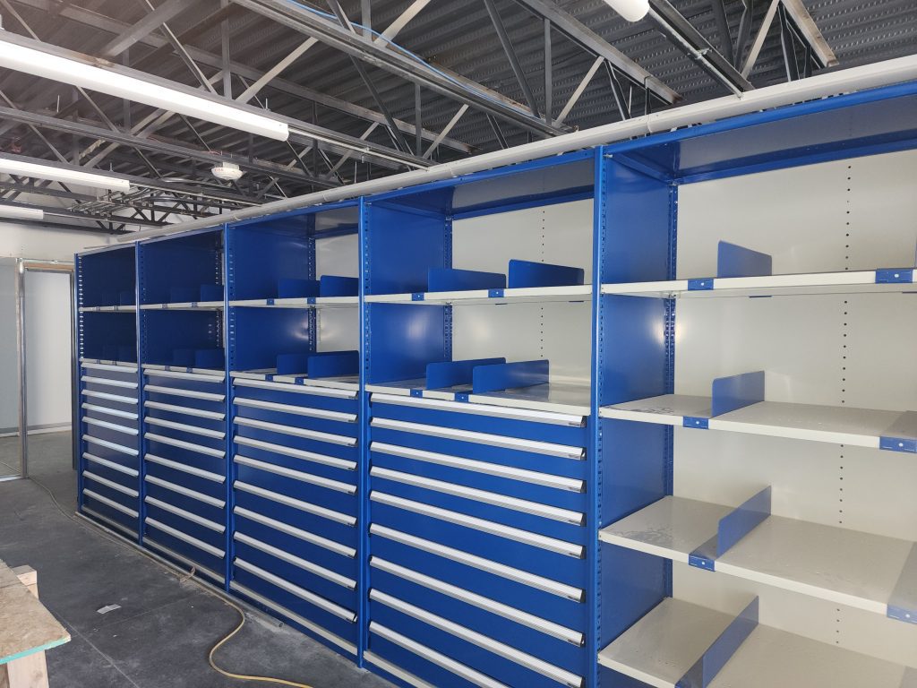 Rows of blue metal shelving units with adjustable dividers and drawers, partially assembled, in an industrial or warehouse setting with exposed ceiling beams and unfinished flooring.