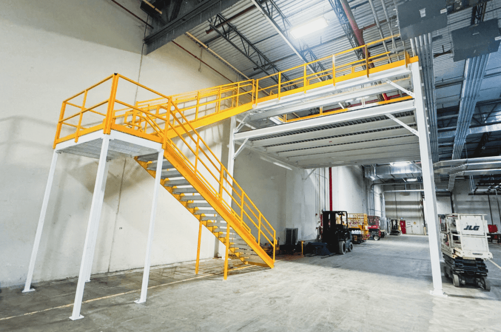 A yellow metal staircase with railings leads to an elevated mezzanine platform inside a spacious industrial warehouse with high ceilings.