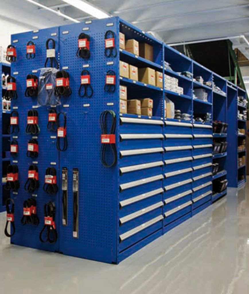 Blue metal shelving units with drawers and pegboards hold various products, showcasing an organized automotive parts room design, with boxed supplies and hanging items neatly arranged in a well-lit industrial storage area.