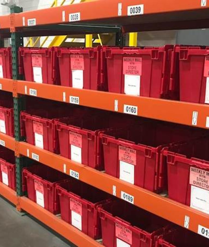 Red plastic bins with white labels showcase a well-organized parts room bin labeling system on orange metal shelves. Each bin is clearly marked with numbers and names for easy identification in the storage area.